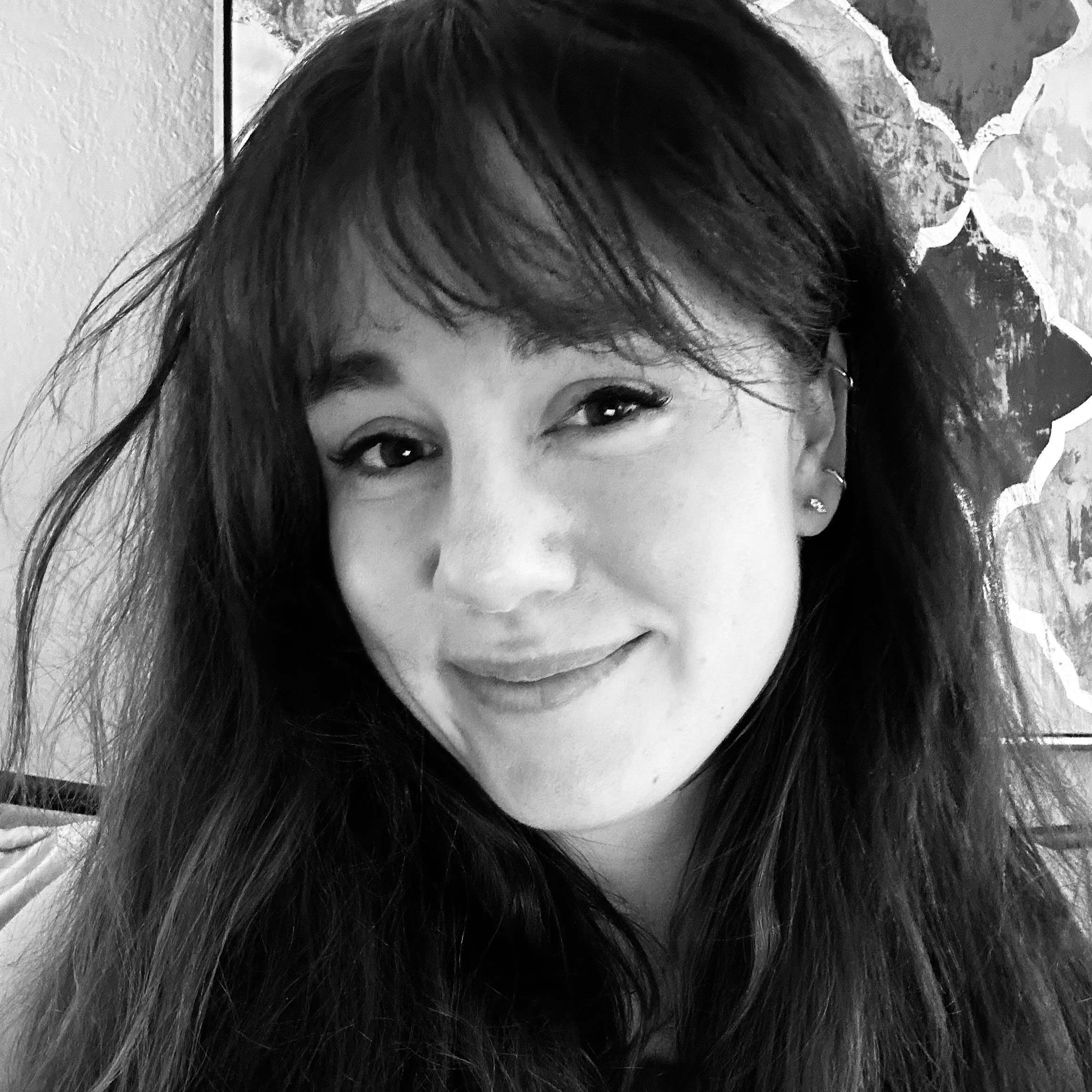 A person with long dark hair and bangs smiles at the camera. They are wearing stud earrings and are in front of a framed abstract artwork. The image is in black and white.